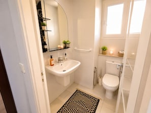 A bathroom with a white sink, toilet, and shower. There is a mirror above the sink, a shelf with toiletries, and a small window. - CosyStay Apartment (Tunis)
