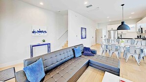 Un salon moderne avec un canapé d’angle, un coin repas avec des chaises, une cuisine en arrière-plan et des œuvres d’art accrochées aux murs.