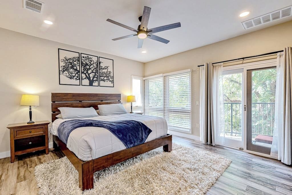 Un dormitorio con cama, ventilador de techo, mesita de noche, lámpara, ventana grande con persianas y vistas al exterior.