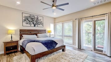 Un dormitorio con cama, ventilador de techo, mesita de noche, lámpara, ventana grande con persianas y vistas al exterior.
