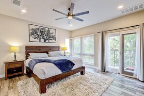 Ein Schlafzimmer mit einem Bett, einem Deckenventilator, einem Nachttisch, einer Lampe, einem großen Fenster mit Jalousien und Blick ins Freie.