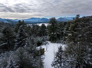 A snowy forest with a cabin, mountains in the background, and a cloudy sky. - Mirrored Glass Cabin with Firepit & Forest Views in Au Sable Forks, New York (Jay)