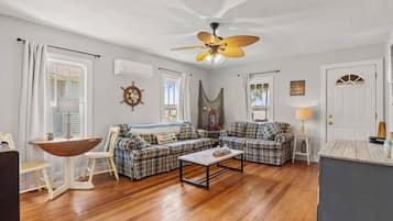 A living room with a wooden floor, two plaid sofas, a coffee table, a ceiling fan, and a door with a sign.