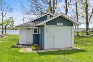 A small, single-story building with a white door and a garage door, labeled 'The Shack,' situated on a grassy area near a body of water.