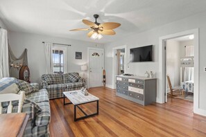 A living room with a plaid sofa, a coffee table, a ceiling fan, a TV, and a wooden floor.