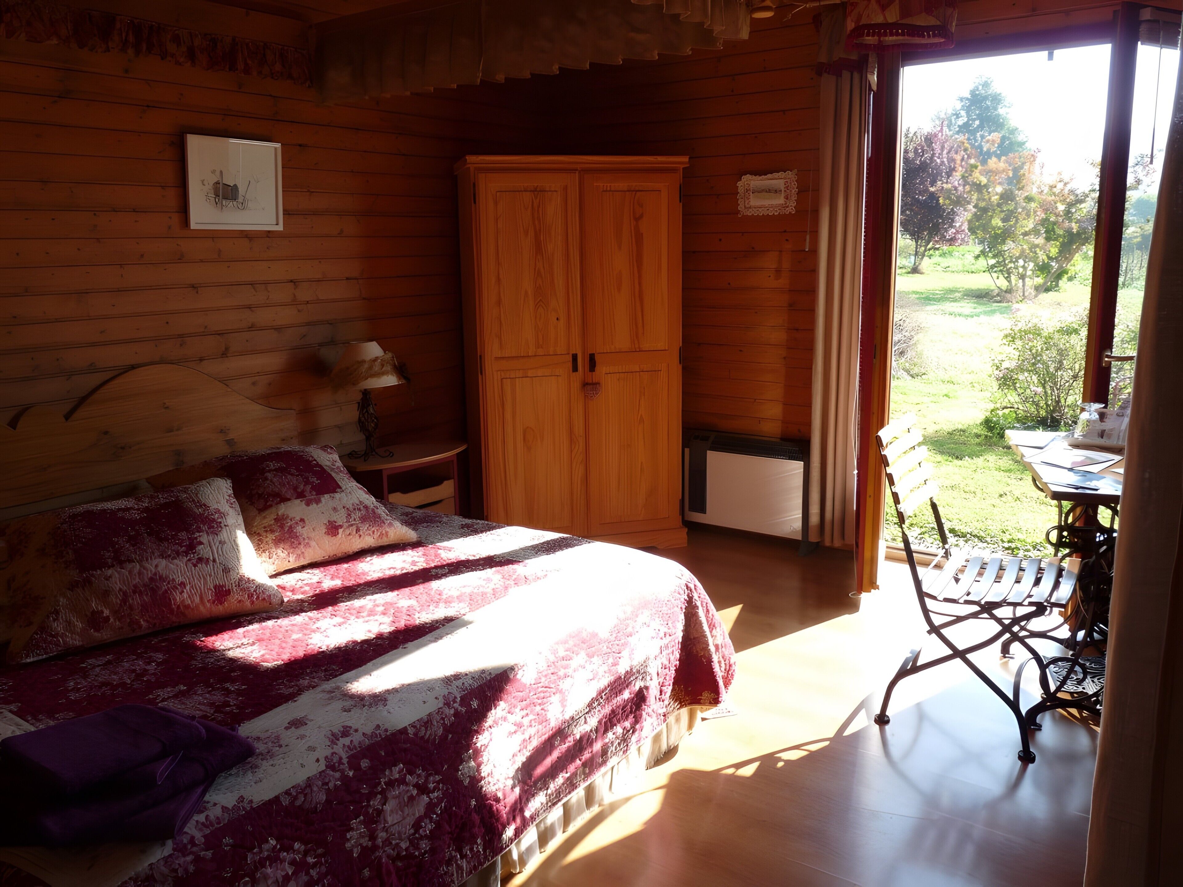 Ein Schlafzimmer einer Holzhütte mit Bett, Kleiderschrank, Tisch und Blick ins Freie.