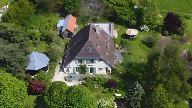 Une vue aérienne d’une maison entourée d’une végétation luxuriante, avec un toit en pente et un jardin bien entretenu.