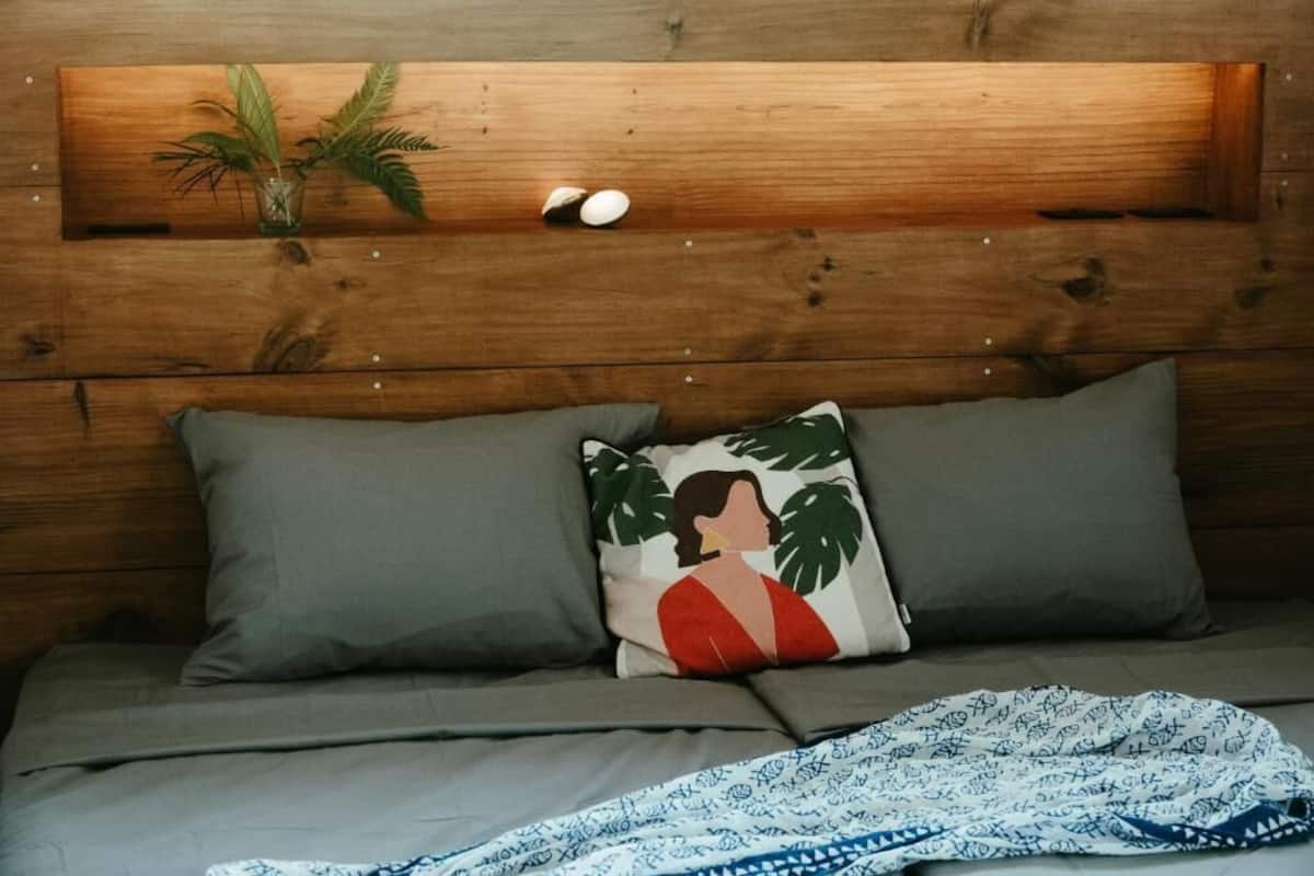 Una cama con ropa de cama gris, un almohadón con diseño de planta y un cabecero de madera con una planta y dos huevos.