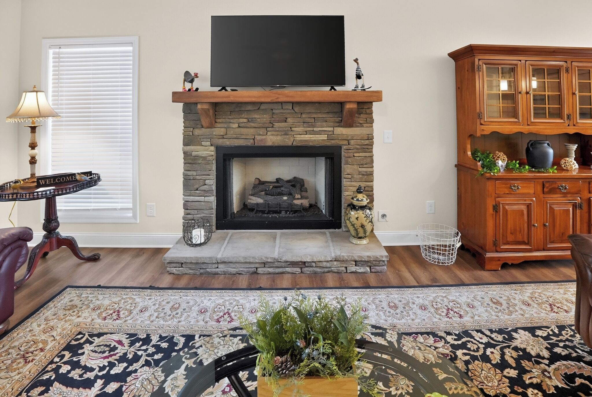Una sala de estar con chimenea de piedra, un televisor de pantalla plana montado encima, un aparador de madera con puertas de cristal y una alfombra estampada.