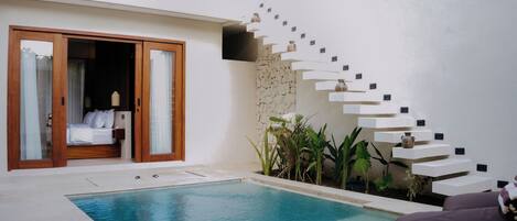 A modern pool area with a clear blue pool, a wooden door leading to a bedroom, and a unique white staircase with shelves.