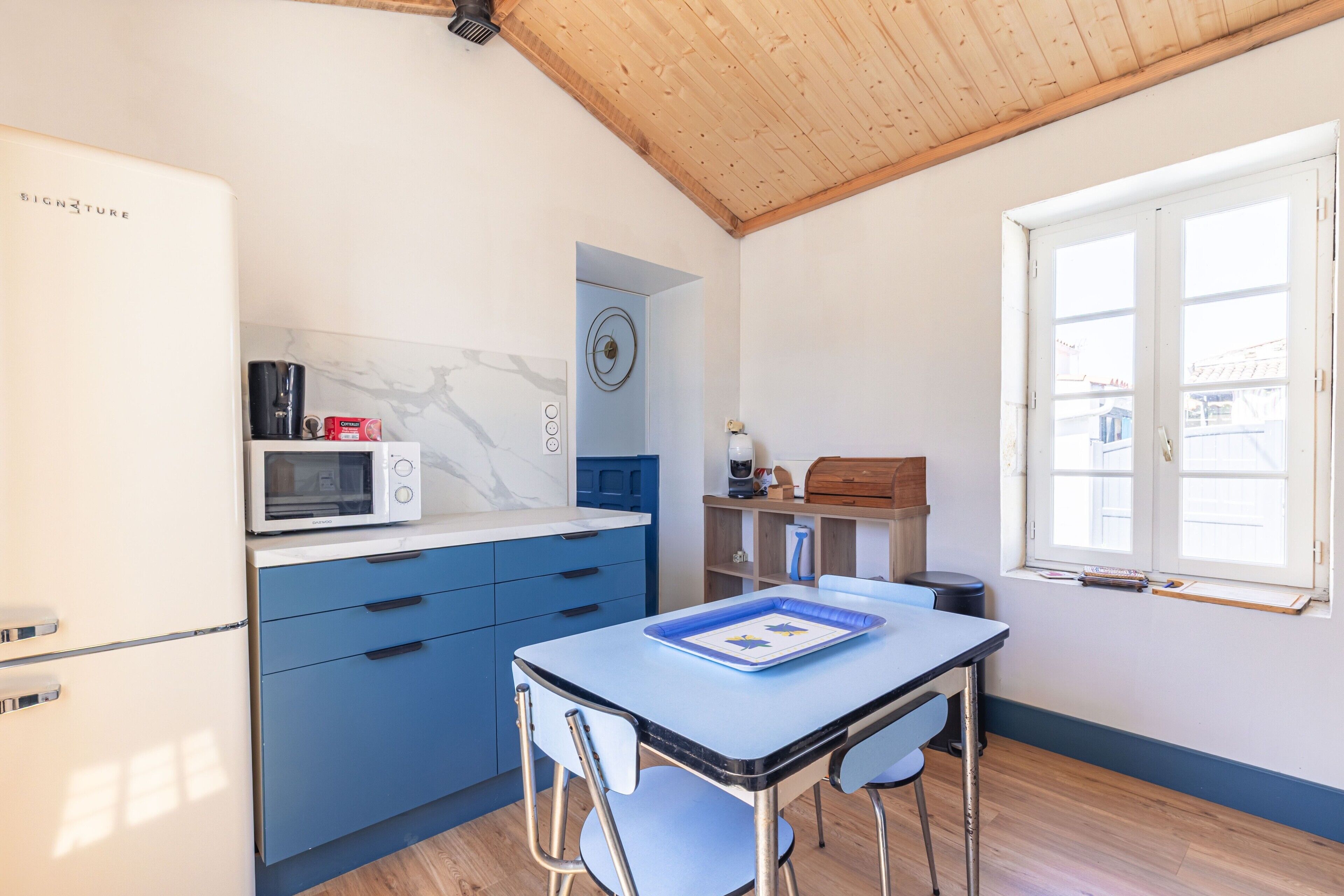 A compact kitchen with a refrigerator, microwave, and blue cabinets. There's a window with a view of buildings outside.