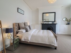 Une chambre à coucher avec un lit, une table de chevet, une lampe, un miroir, une cheminée et un panneau décoratif représentant le mot « AMOUR » accroché au mur.