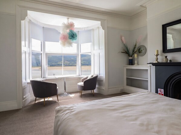 Une chambre avec un grand lit, deux fauteuils, une cheminée, une baie vitrée donnant sur un beau paysage et un miroir au mur.