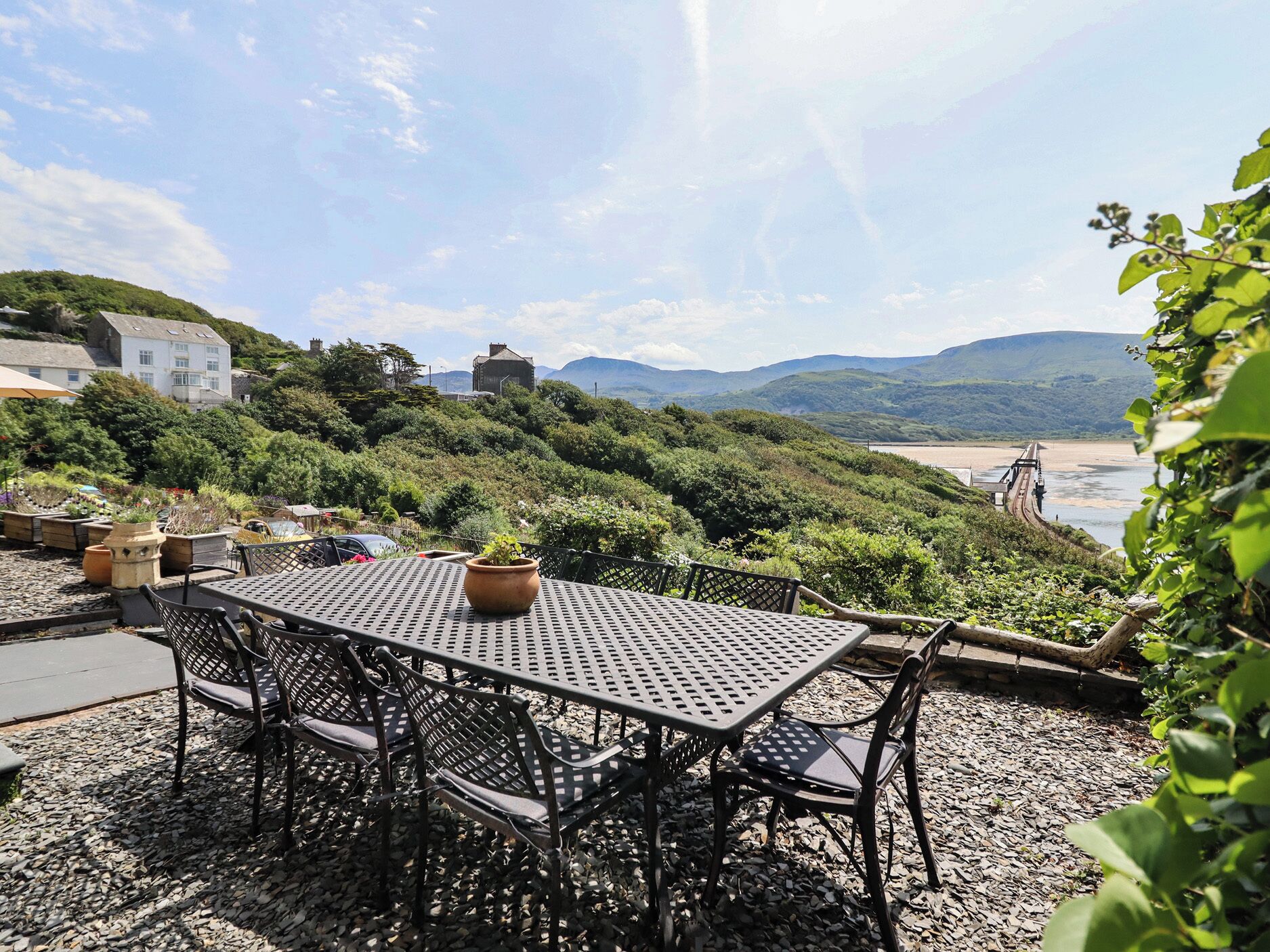 Et bord og stole på en grusbelagt terrasse med udsigt til et kystlandskab og bakker.