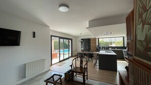Ein geräumiger Wohnbereich mit Esstisch, Stühlen, einer Küche mit großem Fenster und einem Pool, der durch die Glastür zu sehen ist.