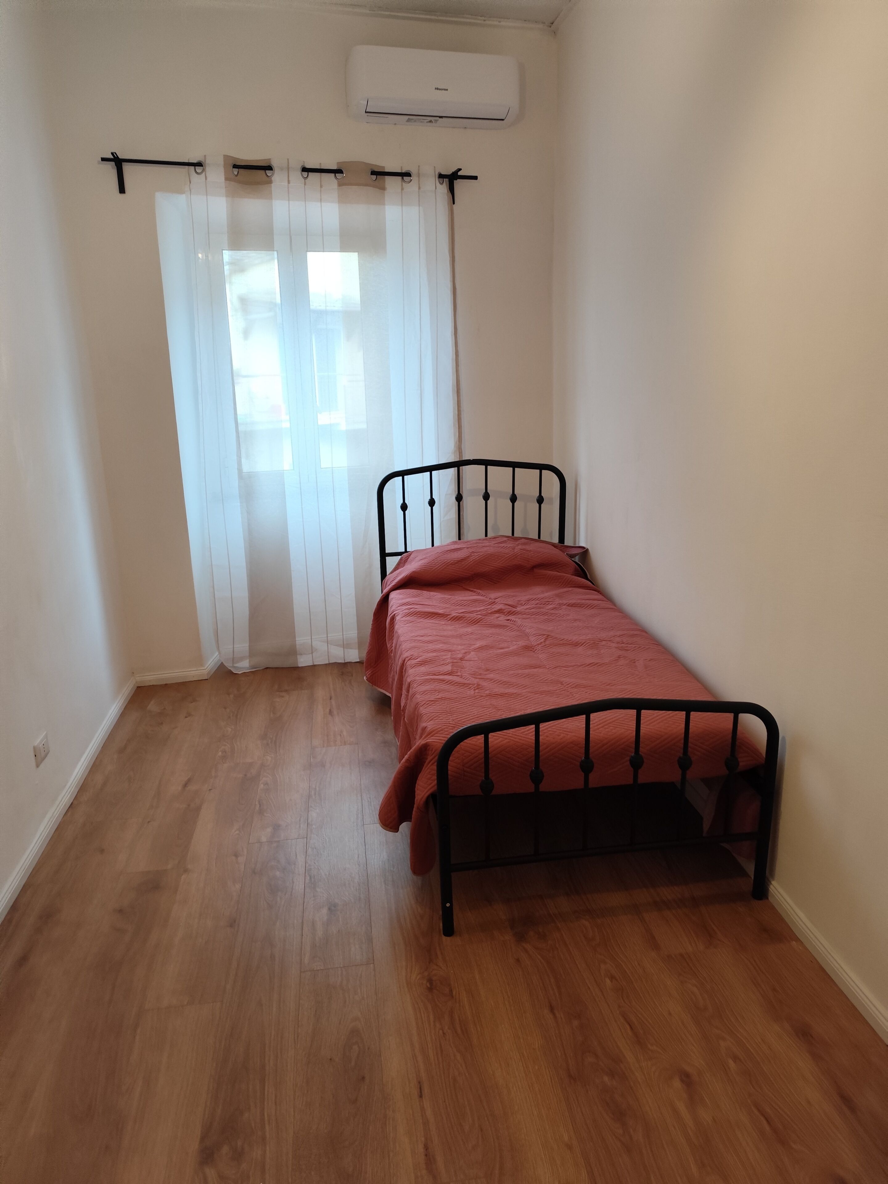 Een kleine kamer met een enkel bed, een raam met dunne gordijnen en een airconditioning op de muur.