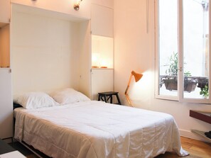 Ein Schlafzimmer mit Bett, Schreibtisch, Stuhl, Lampe und einem Fenster mit Pflanzen.