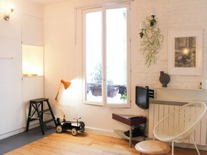 Ein Zimmer mit Holzboden, einem weißen Stuhl, einem kleinen Tisch, einer Lampe, einem Fernseher und einem Fenster mit Blick auf Grün.