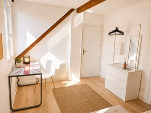 Ein Zimmer mit sichtbarem Holzbalken, einem weißen Schminktisch, einem Spiegel, einem schwarzen Kleiderbügel, einem Glastisch mit Stuhl, einer kleinen Pflanze und einer brennenden Kerze.