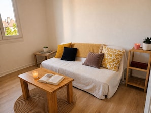 Ein gemütliches Zimmer mit einer Couch, einem kleinen Tisch mit einem Buch, einer Topfpflanze und einem Fenster.