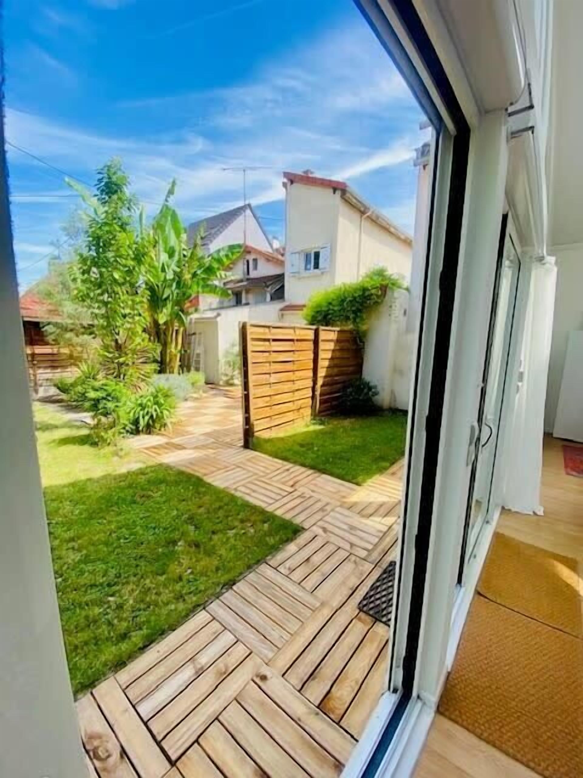 On aperçoit, à travers une porte-fenêtre coulissante, un jardin avec une terrasse en bois, une pelouse et une clôture en bois.