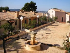 Ein Innenhof mit einem Steinfbrunnen, Gebäuden mit Terrakotta-Dächern und Straßenlaternen.