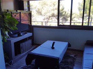 Ein Zimmer mit einem Bett, einem kleinen Tisch, einem Fernseher und einem Fenster mit Blick auf Bäume.
