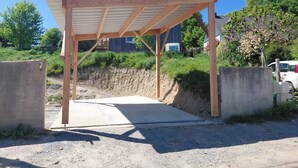 Une structure en bois dotée d’un toit, qui pourrait être un abri de stationnement ou un petit pavillon, entourée d’un mur en béton et bordée d’un chemin de terre.