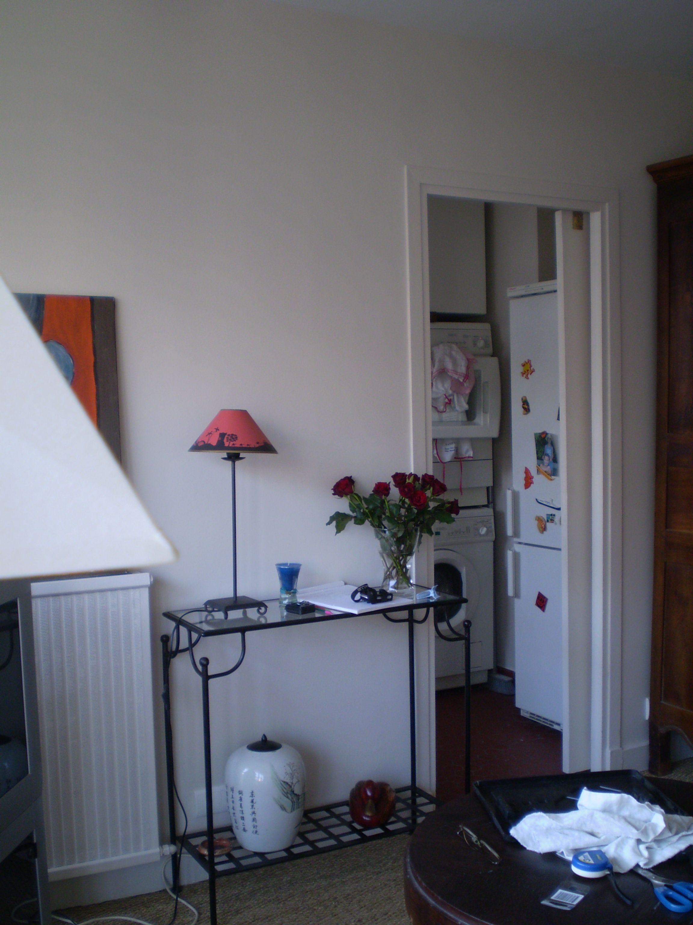 A room with a desk, a lamp, a vase with flowers, a radiator, and a refrigerator.