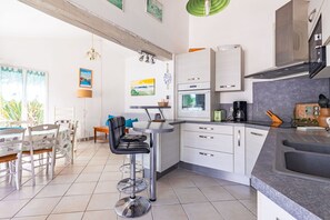 A modern kitchen with white cabinets, a coffee machine, and a dining area with a table and chairs. - Charming Villa “Maison Piscine et Nature” with Private Pool, Terraces, and Garden (Le Château-d'Oléron)