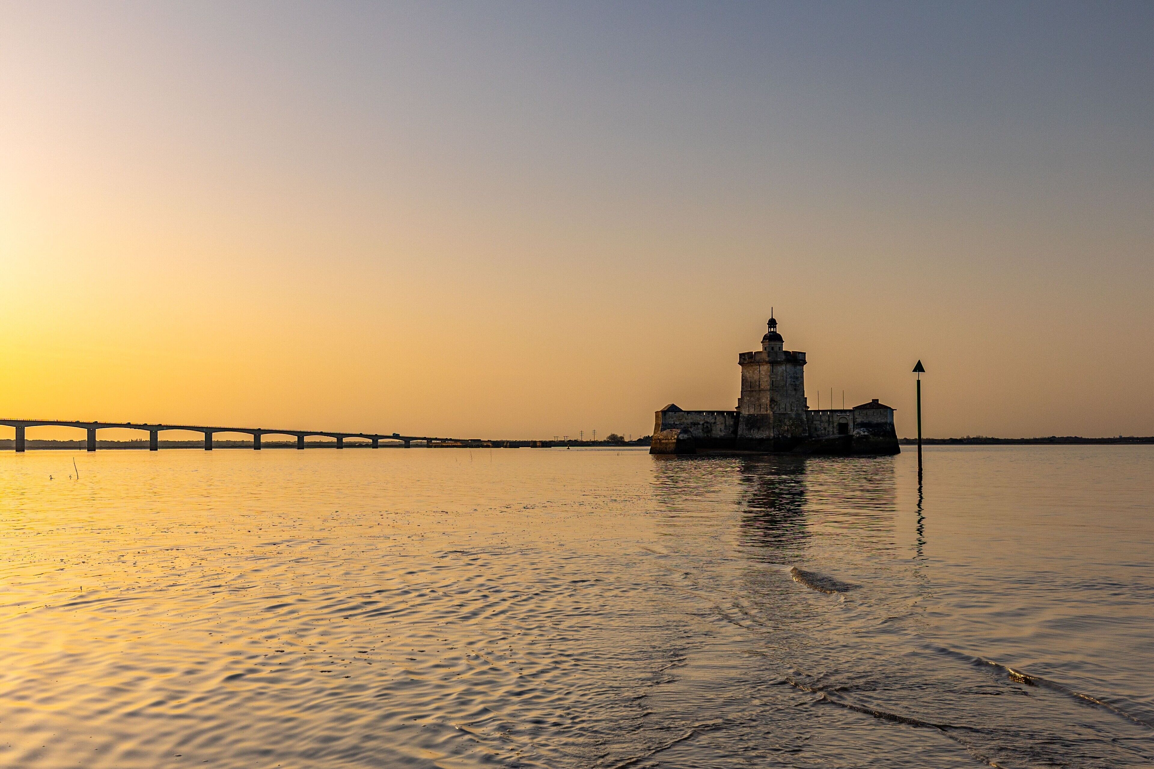 A lighthouse by a calm body of water with a bridge in the background.