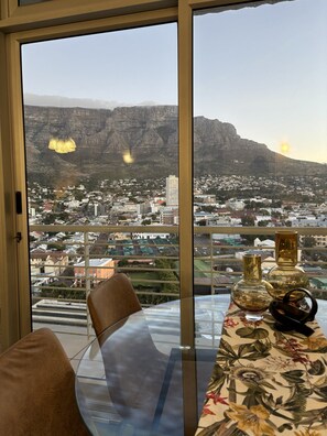 Una habitación con un ventanal grande con vistas a un paisaje urbano y montañas. Hay una mesa con adornos y una silla.