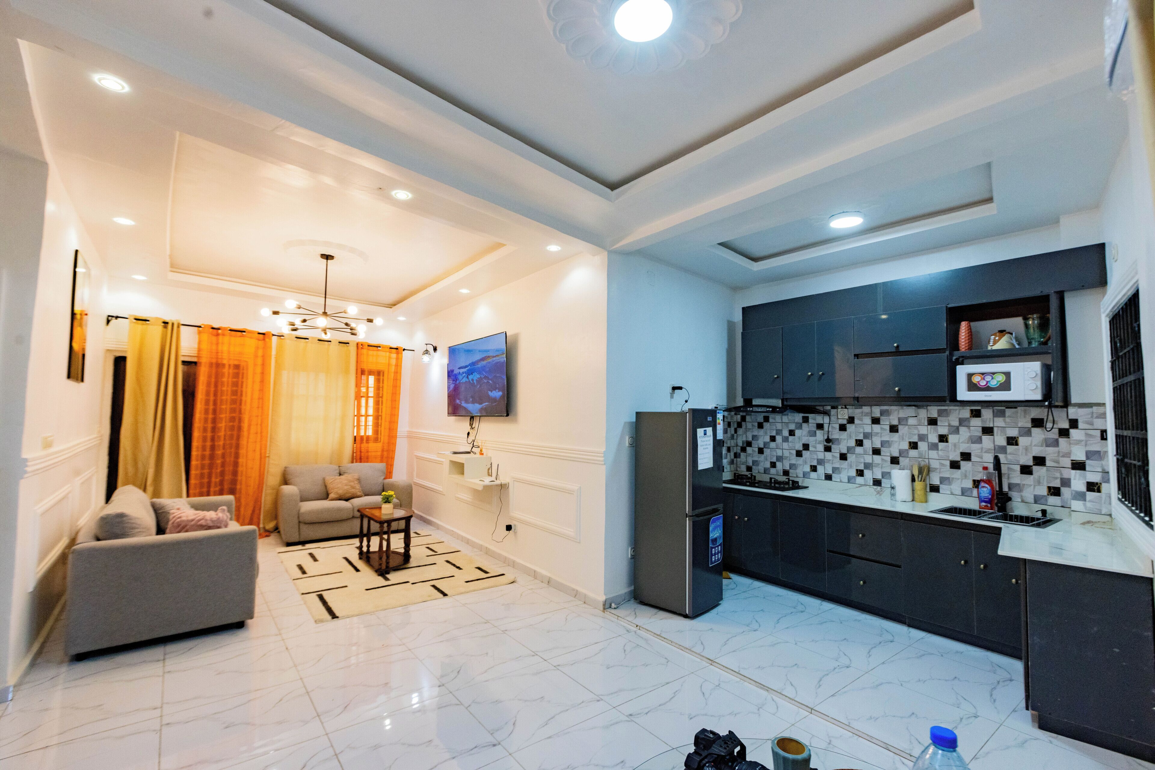Una sala de estar moderna con un sofá, un televisor y una zona de cocina con refrigerador y salpicadero de azulejos.