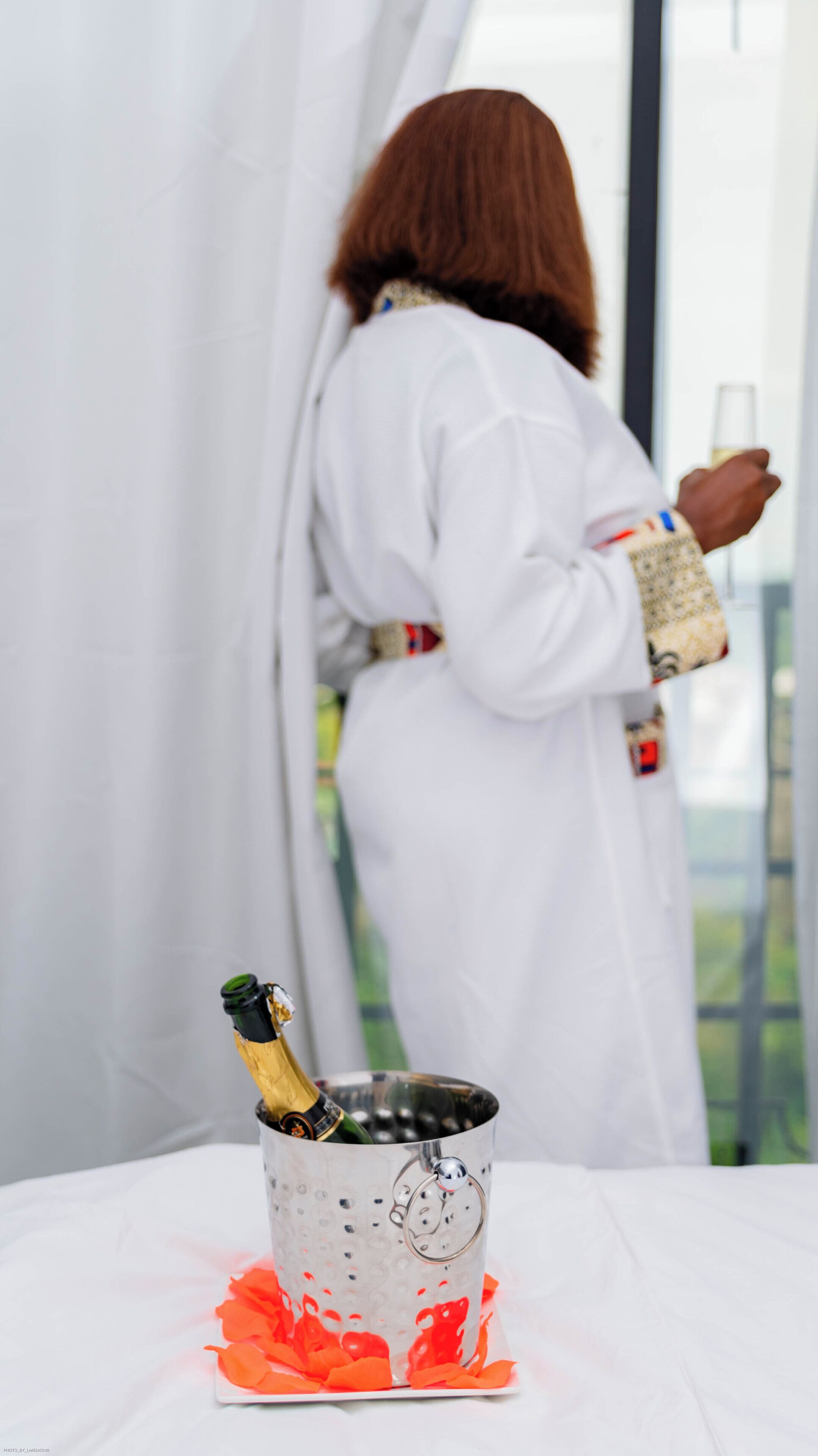 A person in a white robe holding a glass, standing near a table with a champagne bottle and a metal bucket with orange decorations.