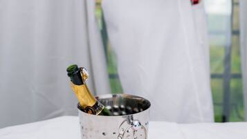 A person in a white robe holding a glass, standing near a table with a champagne bottle and a metal bucket with orange decorations.