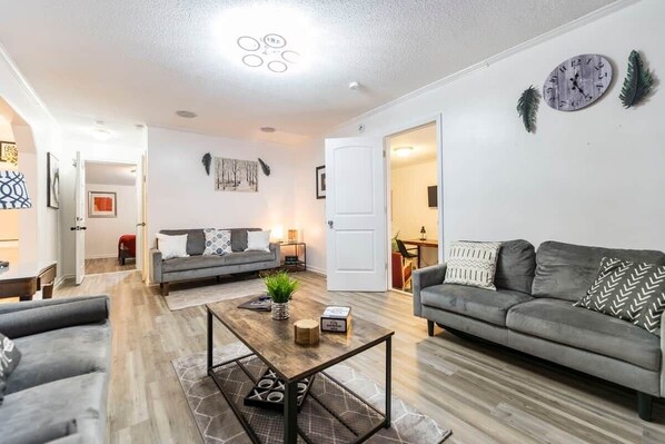 A modern living room with a grey sofa, a coffee table, and a dining area in the background.