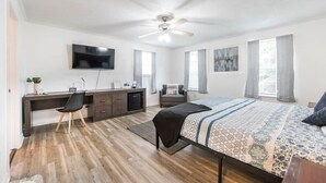 A bedroom with a bed, desk, chair, TV, ceiling fan, and windows.