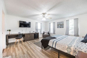 A bedroom with a bed, desk, chair, TV, ceiling fan, and windows.