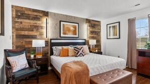 A bedroom with a bed, a chair, a nightstand, a lamp, and a window.
