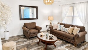 Ein Wohnzimmer mit braunem Sofa, einem runden Couchtisch, einem Sessel, einer Lampe, einem Topfpflanzen und einem Spiegel an der Wand.