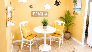 Ein Zimmer mit einer gelben Wand, einem runden Tisch mit zwei Stühlen, einem Schild mit der Aufschrift 'BEACH' und einem Strandbild.