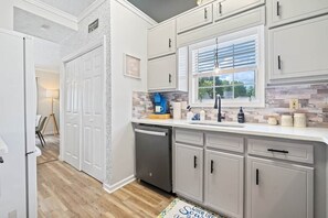 A kitchen with white cabinets, a window with blinds, a dishwasher, and a countertop with various items. - Two Homes in One · 5BR/4BA · Theater & Game (Gwinnett County)
