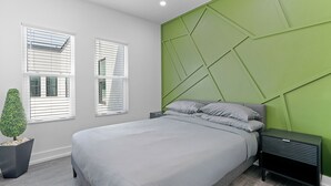 A modern bedroom with a bed, nightstands, a green accent wall, and two windows with blinds.