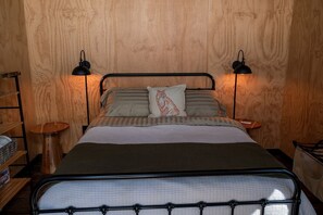 Ein Bett mit Metallgestell, zwei Nachttischlampen, ein Kopfkissen mit rotem Muster und eine Holzwand.