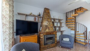 Un soggiorno con camino, televisione, due poltrone, una scala in legno ed elementi decorativi sugli scaffali.