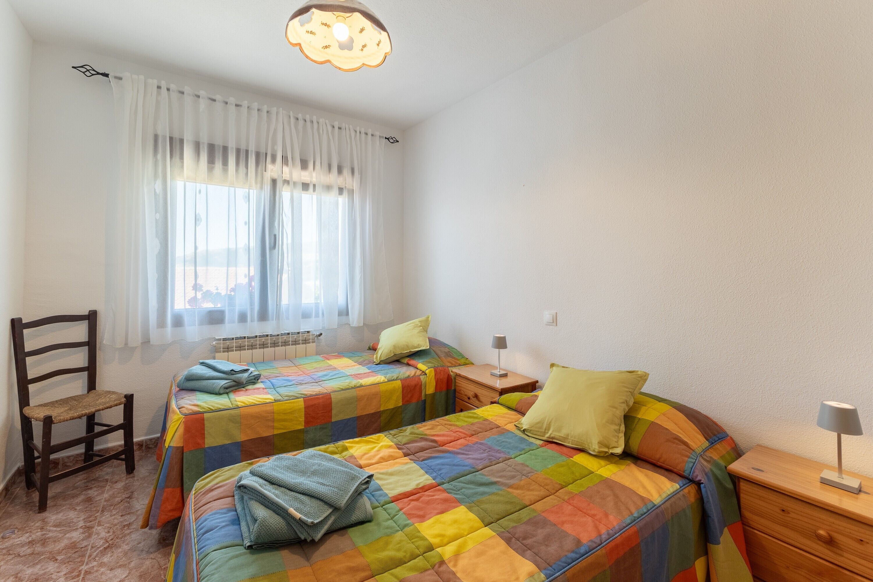 Ein Schlafzimmer mit zwei Betten, einem Sessel, einem Nachttisch, einer Lampe und einem Fenster mit Vorhängen.