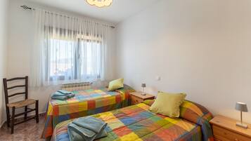 Ein Schlafzimmer mit zwei Betten, einem Sessel, einem Nachttisch, einer Lampe und einem Fenster mit Vorhängen.