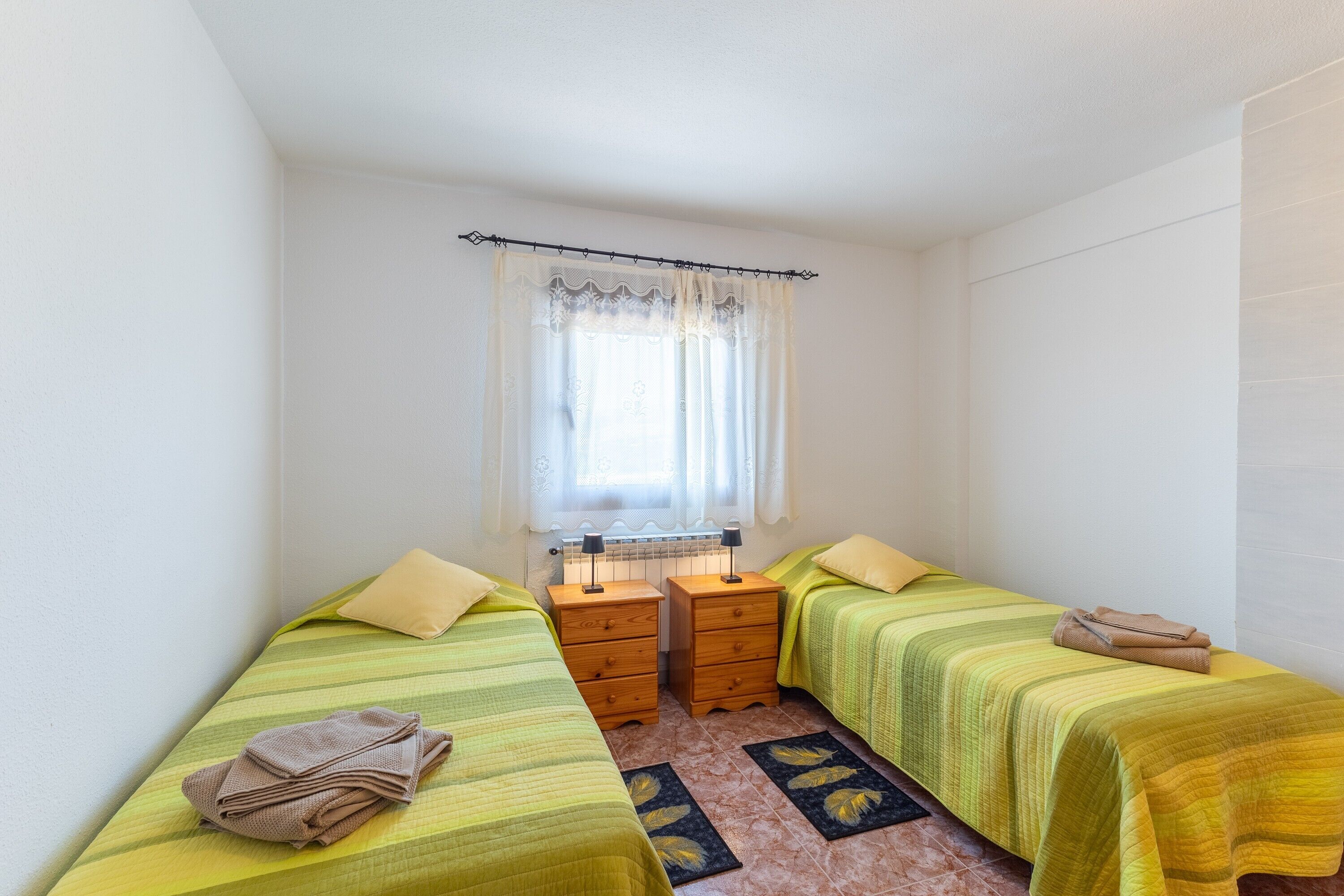 Une petite chambre propre, avec deux lits simples, une fenêtre avec des rideaux, un radiateur et une petite table avec une lampe.