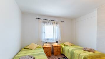 Une petite chambre propre, avec deux lits simples, une fenêtre avec des rideaux, un radiateur et une petite table avec une lampe.