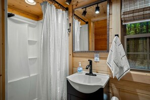 A wooden cabin bathroom with a shower, sink, mirror, and window. - Escape to a Charming Cabin with Hot Tub & Covered Porch in Mentone, Alabama (Mentone)